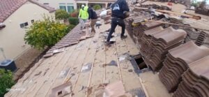PHOTO-2025-01-30-06-50-06 - Desert Sands Contracting Construction workers wearing safety gear on a damaged clay tile roof, removing debris as stacks of old tiles lie to the right.