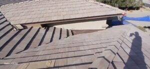 PHOTO-2025-02-01-15-52-42 - Desert Sands Contracting Aerial view of a house roof with gray tile shingles, under construction; blue tarp, ladder visible in the background and the photographer's shadow on the tiles.
