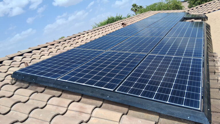 Solar panels mounted on a brown tiled roof with a safety mesh along the edge and blue sky in the background.