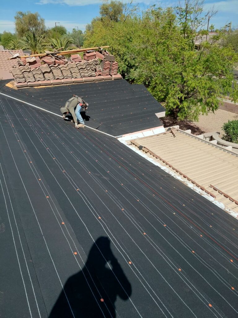 Roofing worker kneels on a dark metal roof, installing underlayment with orange fasteners; tile stacks sit on the ridge behind.