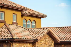 Slates Roof Home Roof - Desert Sands Contracting - Desert Sands Contracting Two-story stucco house with terracotta roof tiles, arched windows, and a stone foundation against a clear blue sky.
