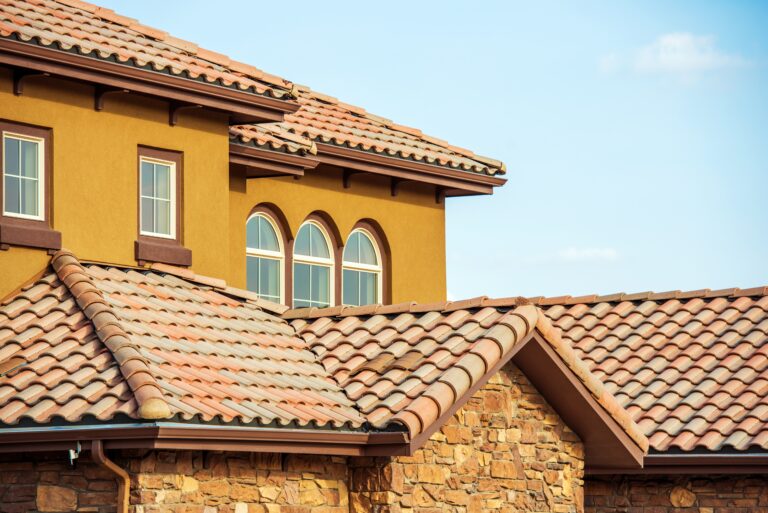 Two-story stucco house with terracotta roof tiles, arched windows, and a stone foundation against a clear blue sky.