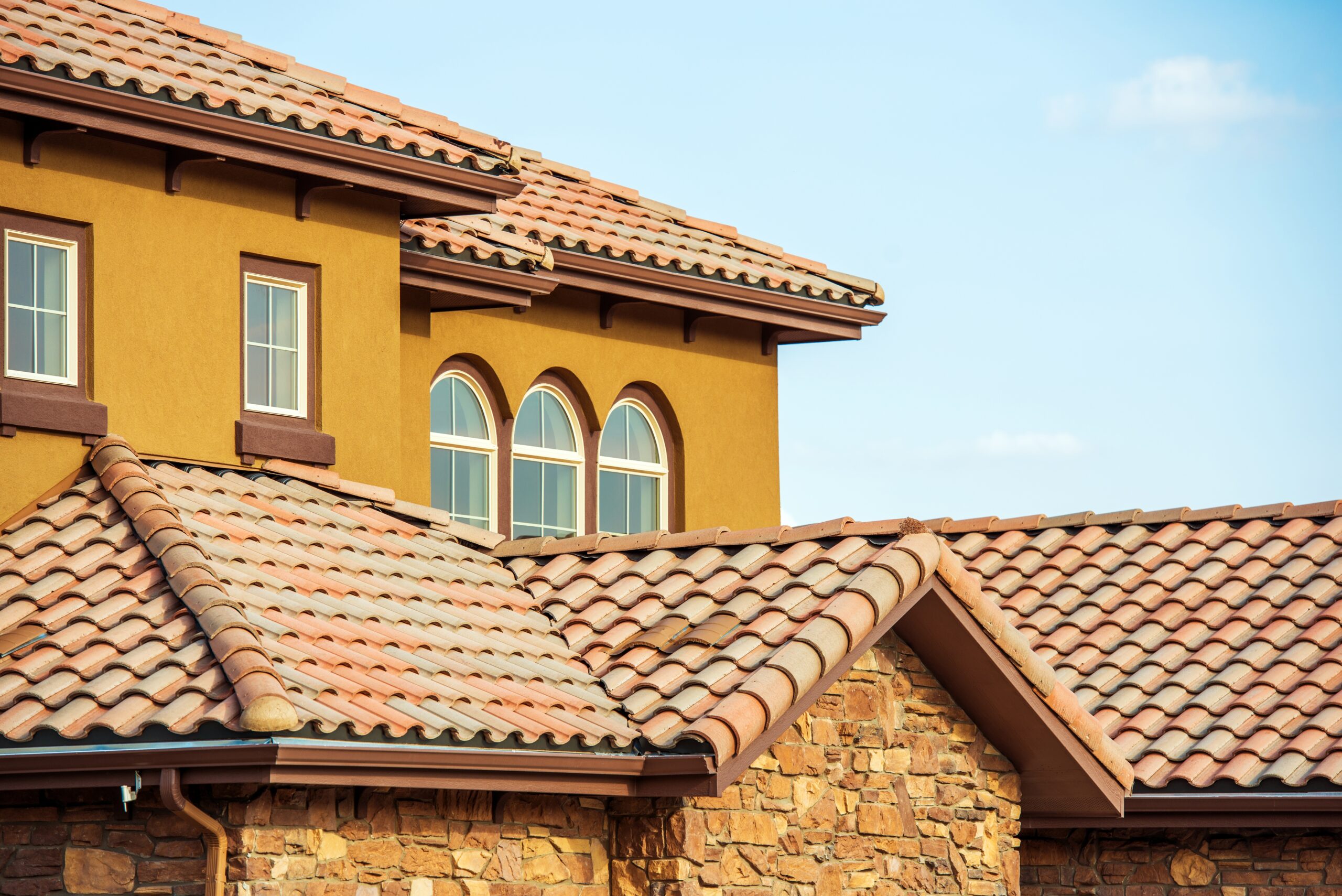 Two-story stucco house with terracotta roof tiles, arched windows, and a stone foundation against a clear blue sky.